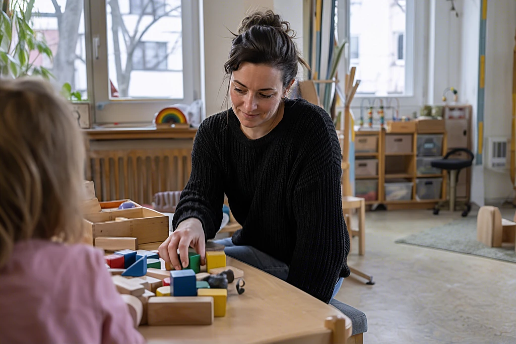 Erzieherin sortiert Bausteine an einem niedrigen Tisch in einem hellen Kita-Gruppenraum